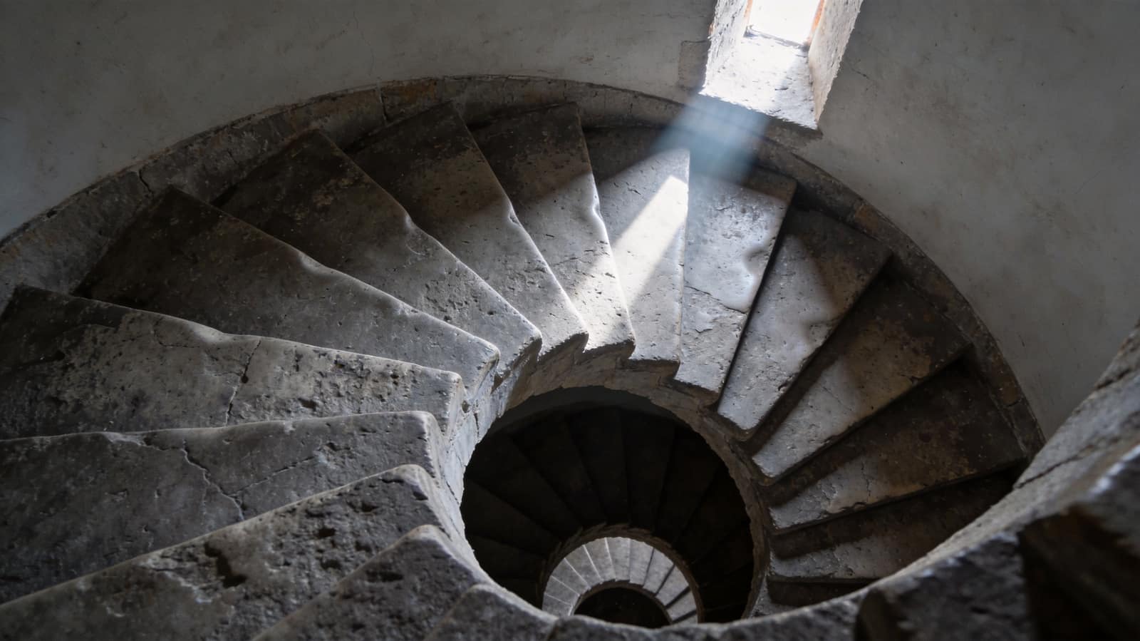 A stone spiral staircase seen from above, each tread dished by use, a single shaft of cold light falling through it