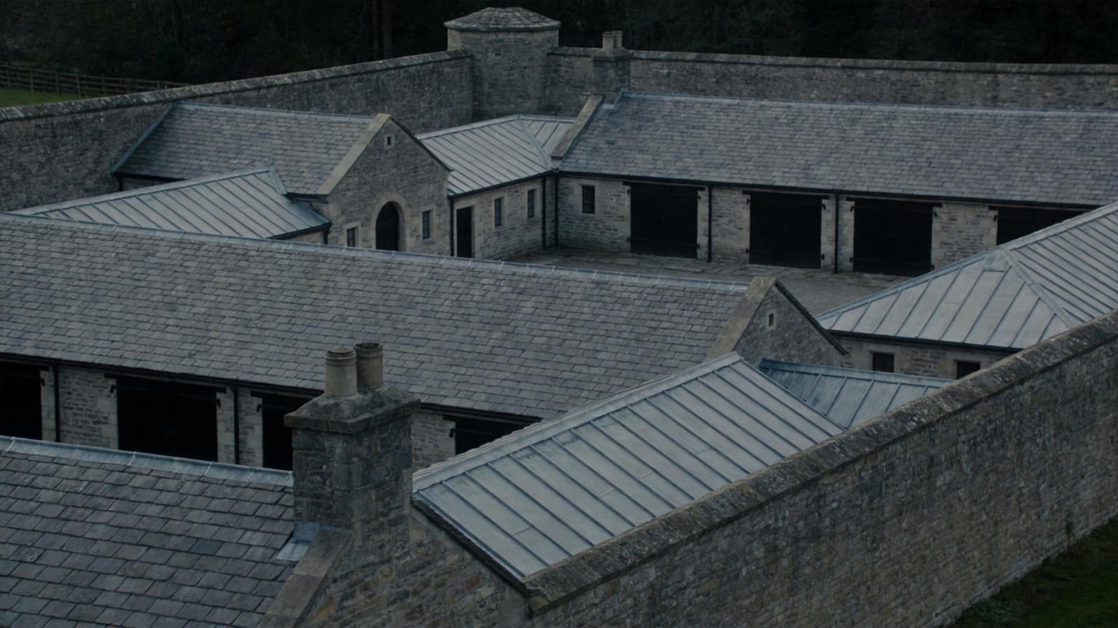 An elevated view across the slate and lead roofs of a walled estate’s outbuildings, many ranges inside one boundary wall