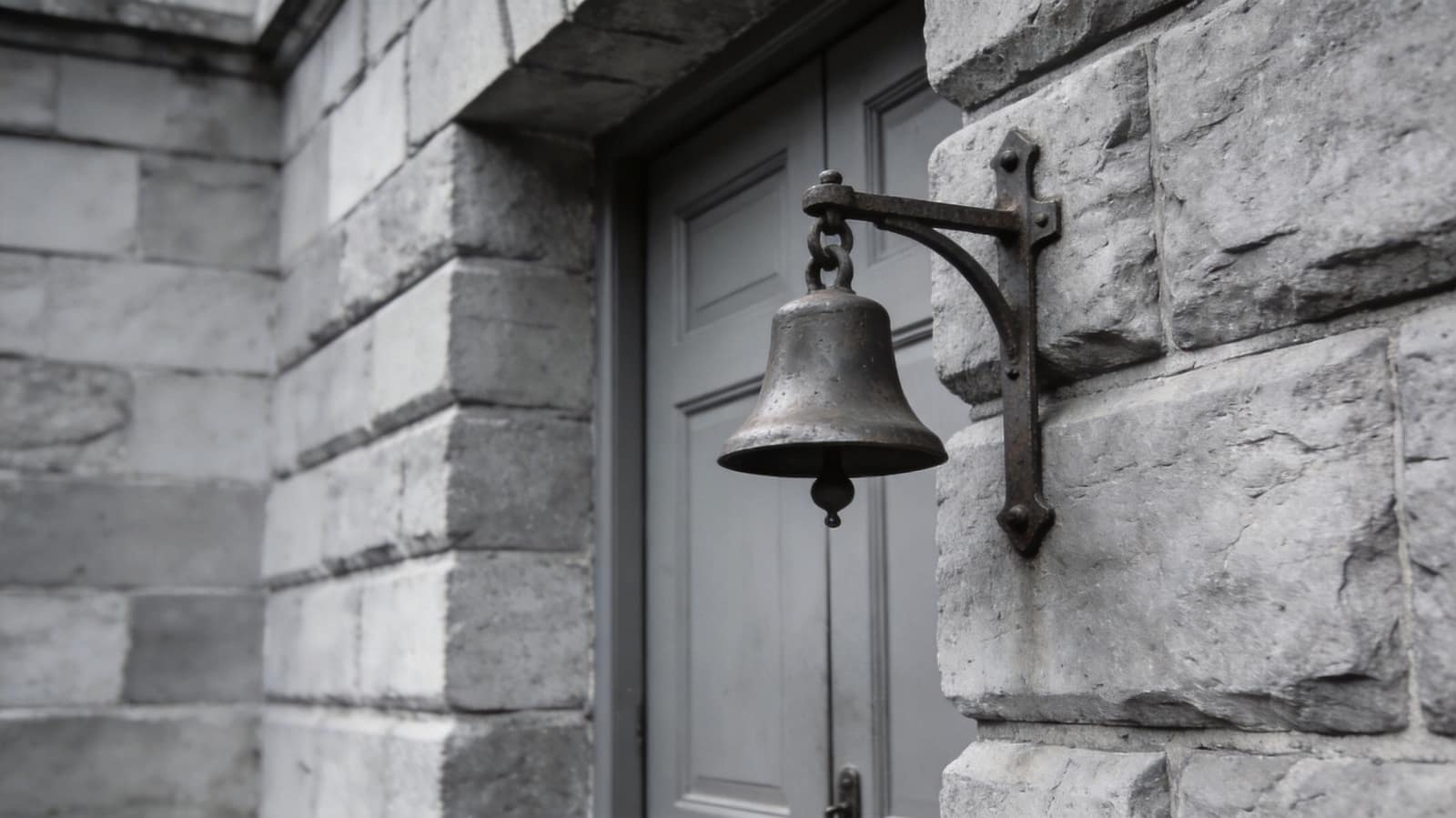 A heavy iron bell mounted on dressed stone beside a closed door, the pull worn smooth and hanging still