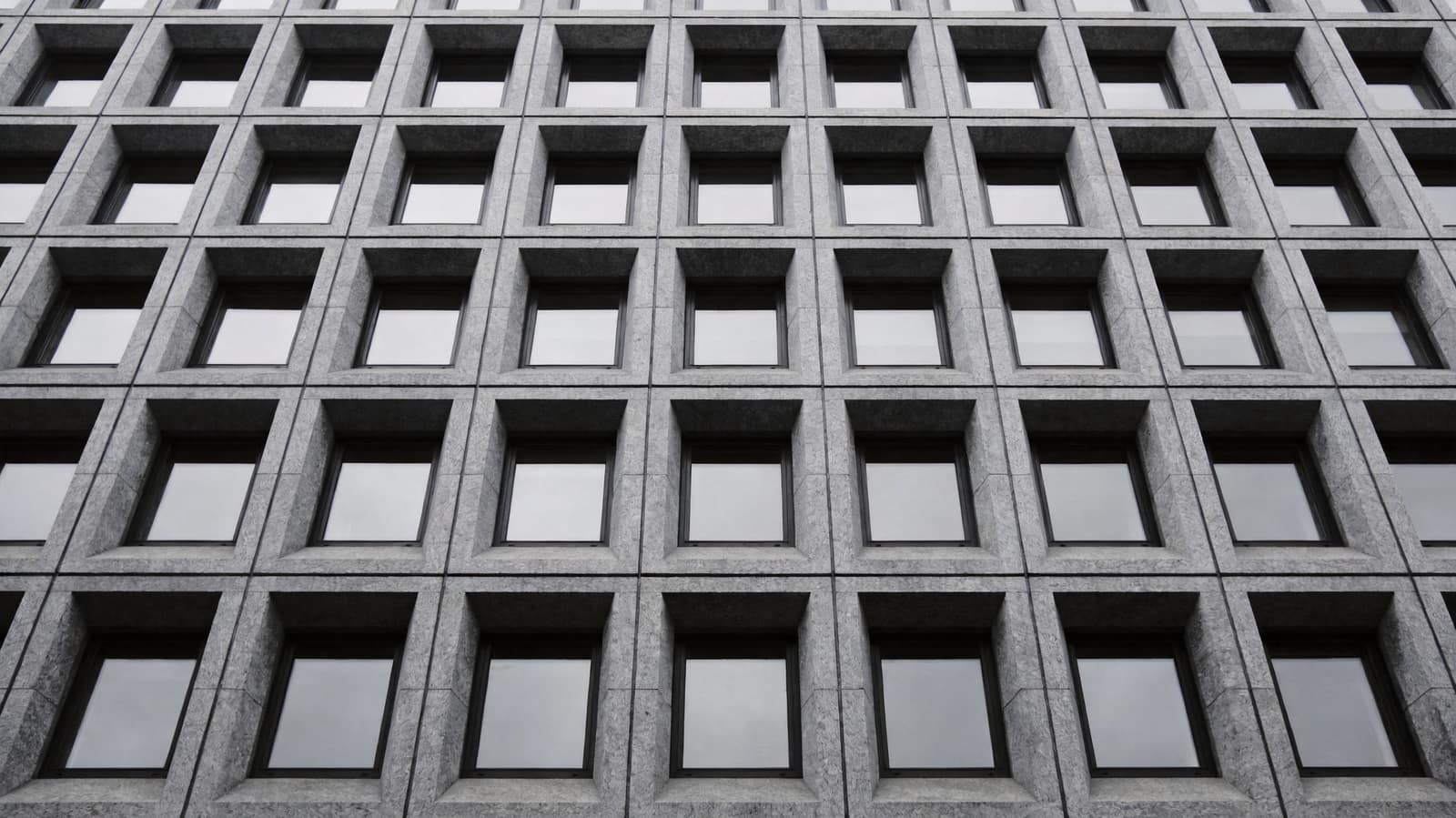 The corner of a severe unadorned ashlar building in flat daylight, its tall windows dark and its facade carrying no signage