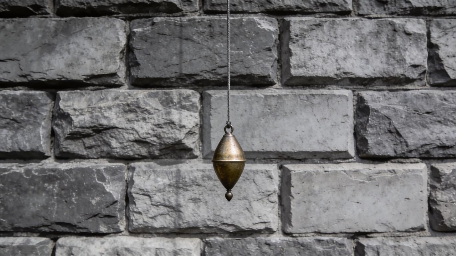 A brass plumb bob hanging dead still on its line against a dressed stone wall