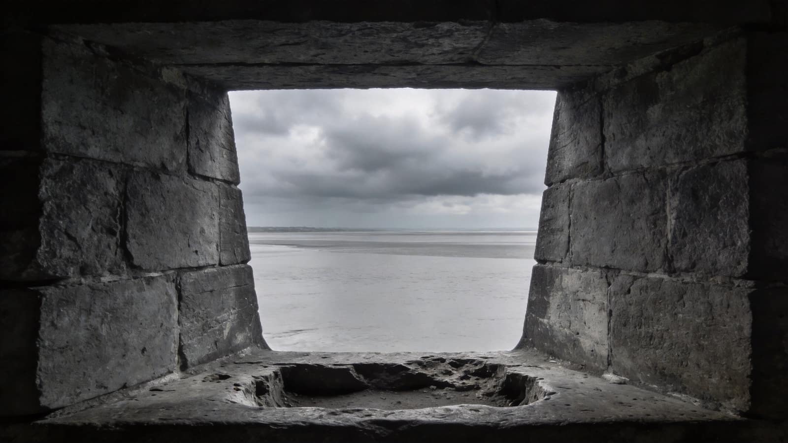The inside of a stone sentry embrasure looking out through a narrow splayed opening onto an empty grey estuary